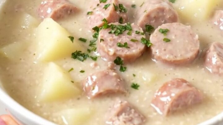A steaming bowl of creamy sausage and potato chowder served in a white bowl with a blurred kitchen background.