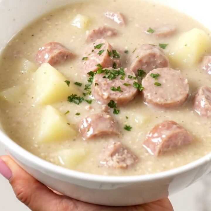 A steaming bowl of creamy sausage and potato chowder served in a white bowl with a blurred kitchen background.