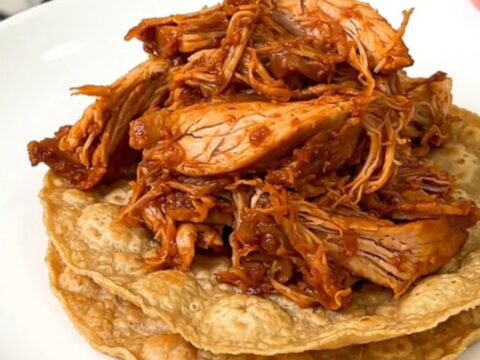 A close-up shot of steaming shredded chicken tinga piled high on crisp tostadas on a white plate.