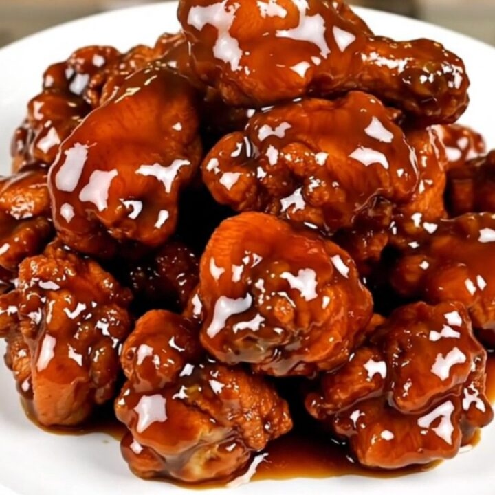 A close-up of glossy General Tso chicken on a white plate, steaming slightly.