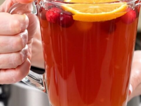 A clear glass mug filled with steaming cranberry apple cider topped with a fresh orange slice.