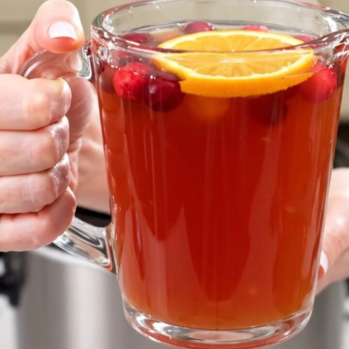 A clear glass mug filled with steaming cranberry apple cider topped with a fresh orange slice.