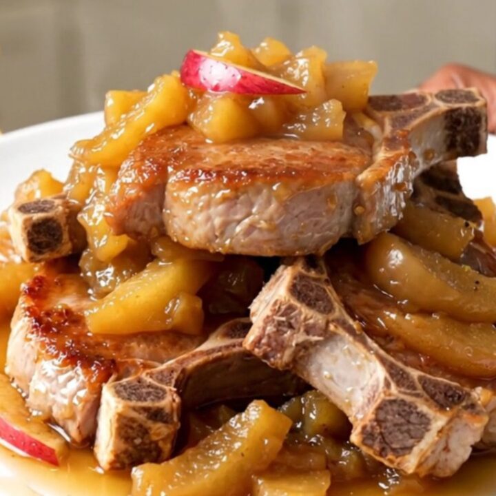 A close-up of tender, slow-cooked pork chops topped with soft, glazed apple slices on a white plate.