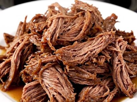 A close-up of a tender, shredded beef chuck roast in a slow cooker with visible onions and a rich, dark gravy.