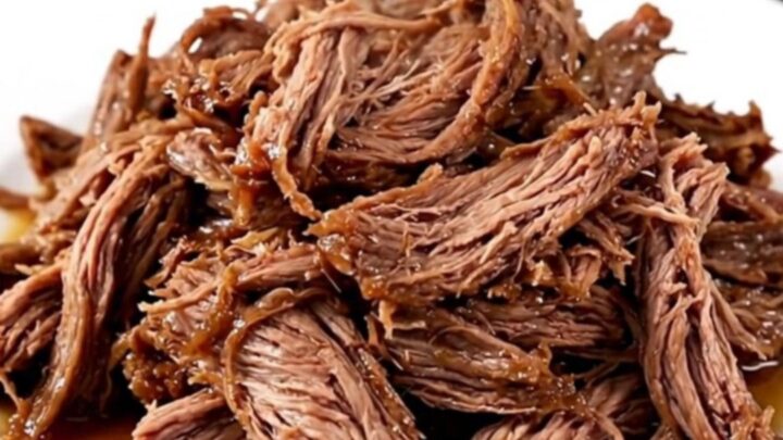 A close-up of a tender, shredded beef chuck roast in a slow cooker with visible onions and a rich, dark gravy.