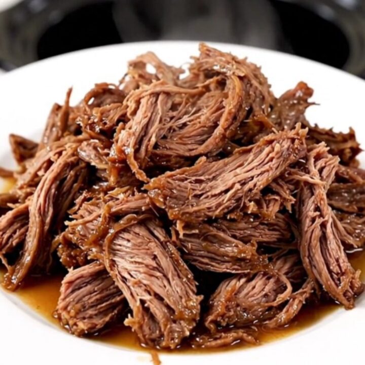 A close-up of a tender, shredded beef chuck roast in a slow cooker with visible onions and a rich, dark gravy.