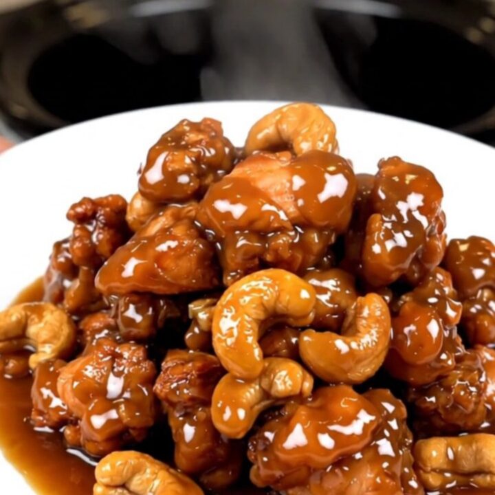 A close-up of glossy cashew chicken in a slow cooker with visible cashews and a rich sauce.