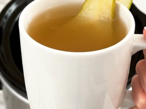 A steaming mug of spiced pear cider garnished with a fresh pear slice, held against a blurred kitchen background.