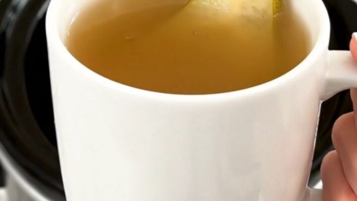 A steaming mug of spiced pear cider garnished with a fresh pear slice, held against a blurred kitchen background.