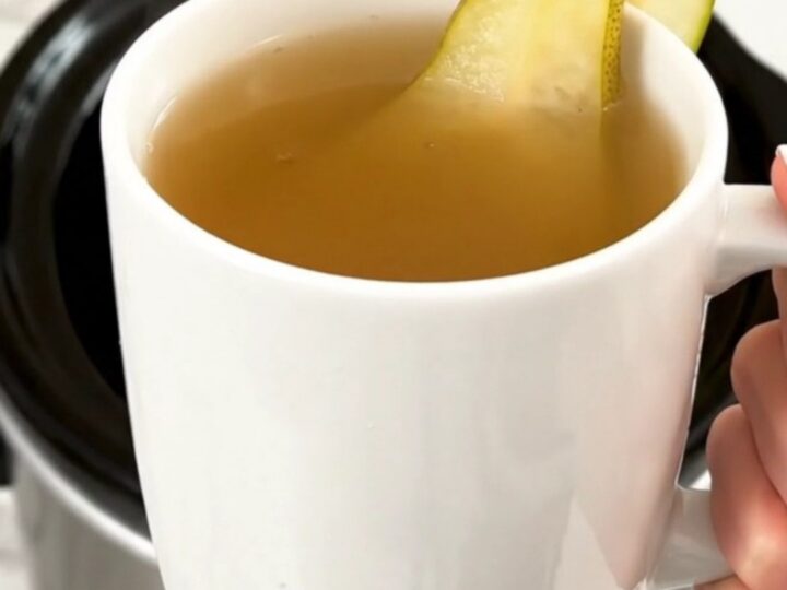 A steaming mug of spiced pear cider garnished with a fresh pear slice, held against a blurred kitchen background.