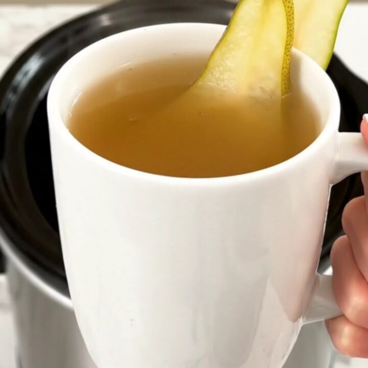 A steaming mug of spiced pear cider garnished with a fresh pear slice, held against a blurred kitchen background.