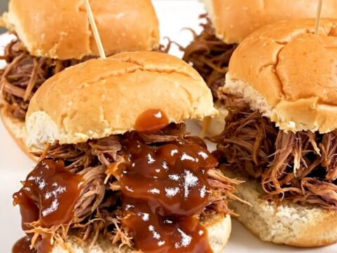 A close-up of juicy, shredded pulled pork on slider buns, steaming on a white plate.