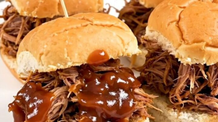 A close-up of juicy, shredded pulled pork on slider buns, steaming on a white plate.