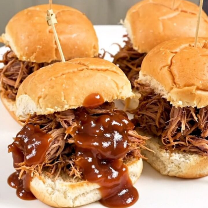 A close-up of juicy, shredded pulled pork on slider buns, steaming on a white plate.