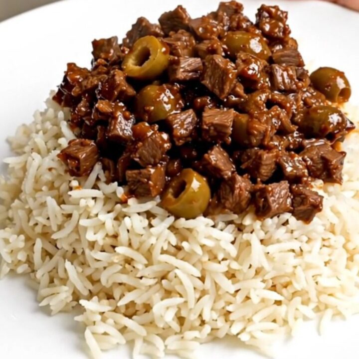 A close-up of a steaming bowl of beef picadillo served over fluffy white rice on a clean white plate.