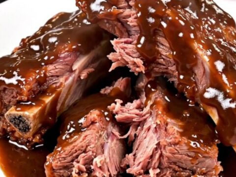 A top-down view of raw beef short ribs, chopped red onion, and barbecue sauce inside a black slow cooker.
