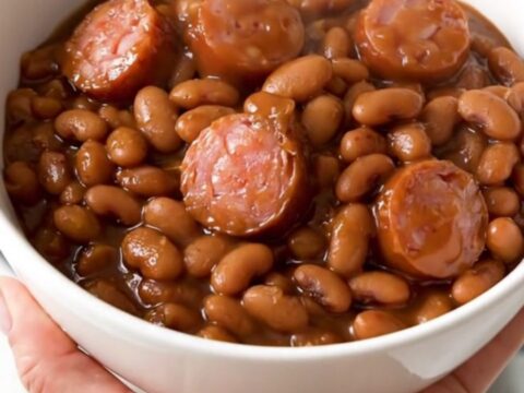 A top-down view of a black slow cooker filled with a rich, glossy mixture of barbecue sausage and baked beans.