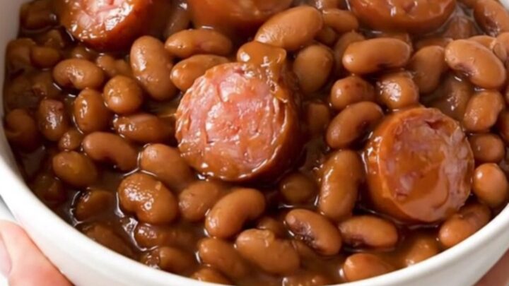 A top-down view of a black slow cooker filled with a rich, glossy mixture of barbecue sausage and baked beans.