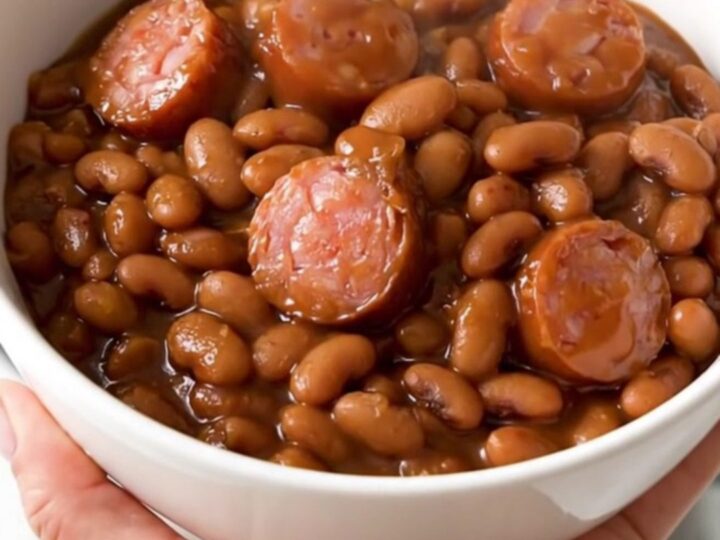 A top-down view of a black slow cooker filled with a rich, glossy mixture of barbecue sausage and baked beans.