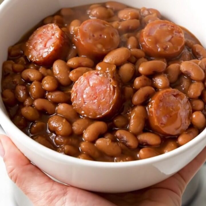 A top-down view of a black slow cooker filled with a rich, glossy mixture of barbecue sausage and baked beans.
