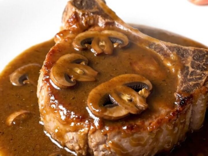 A close-up of a juicy bone-in pork chop topped with creamy mushroom gravy on a white plate.