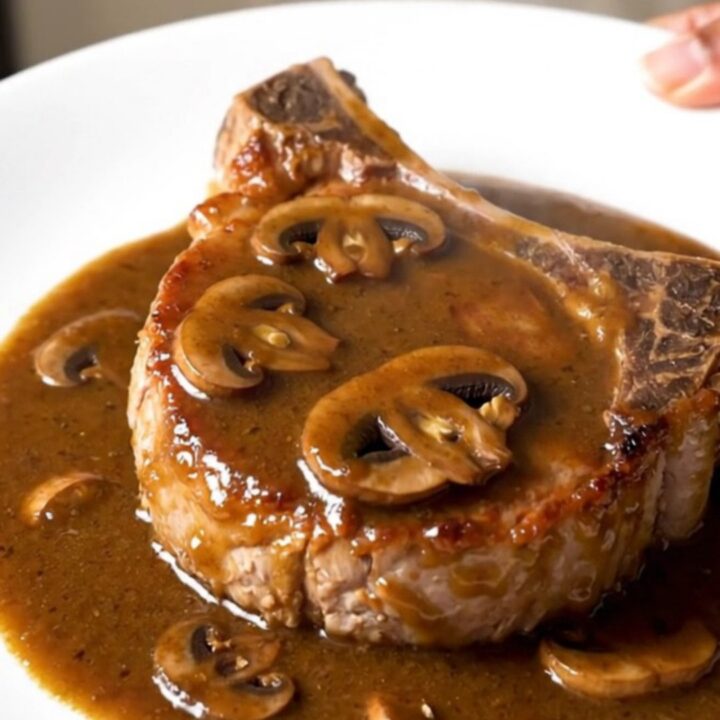 A close-up of a juicy bone-in pork chop topped with creamy mushroom gravy on a white plate.