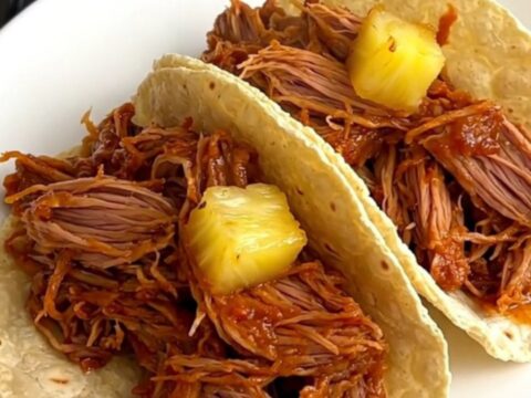 A close-up of tender, shredded al pastor pork with pineapple and red onion, served on warm corn tortillas.