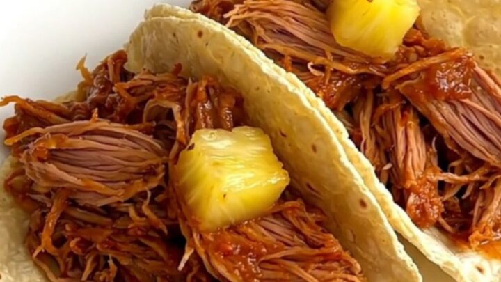 A close-up of tender, shredded al pastor pork with pineapple and red onion, served on warm corn tortillas.