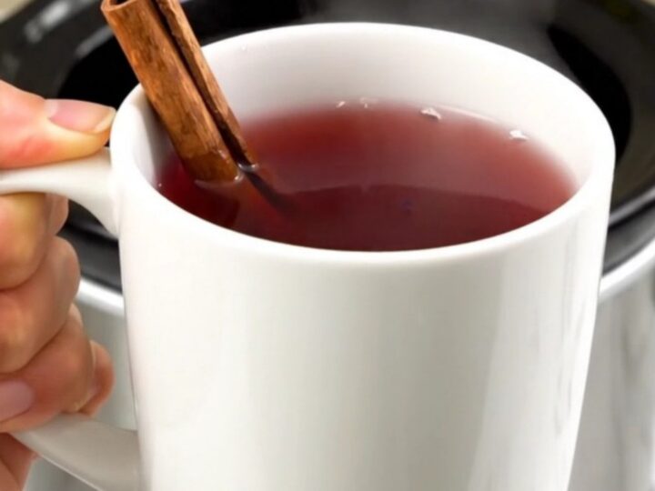 A clear view into a slow cooker filled with cranberry wassail, cinnamon sticks, and spices.