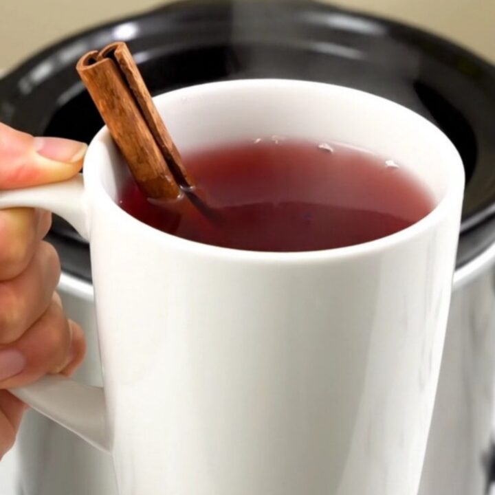A clear view into a slow cooker filled with cranberry wassail, cinnamon sticks, and spices.