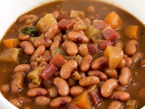 A close-up of a slow cooker filled with a rich, dark pinto bean and bacon stew on a white countertop.