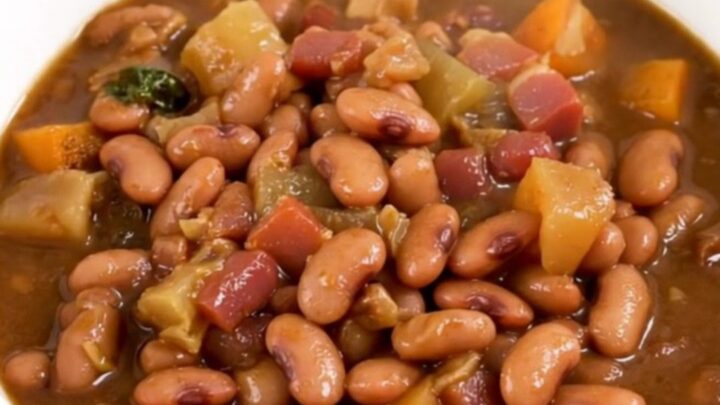 A close-up of a slow cooker filled with a rich, dark pinto bean and bacon stew on a white countertop.