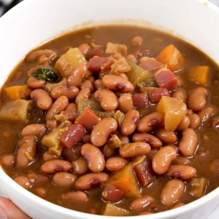 A close-up of a slow cooker filled with a rich, dark pinto bean and bacon stew on a white countertop.