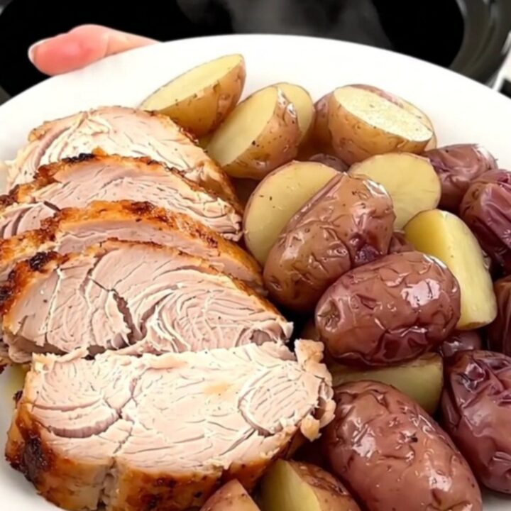 A slow cooker filled with a cooked turkey breast, baby red potatoes, and savory herb broth on a white quartz countertop.