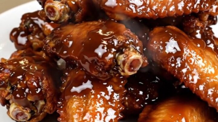 A close-up of glossy, dark-glazed chicken wings in a slow cooker on a white speckled countertop.