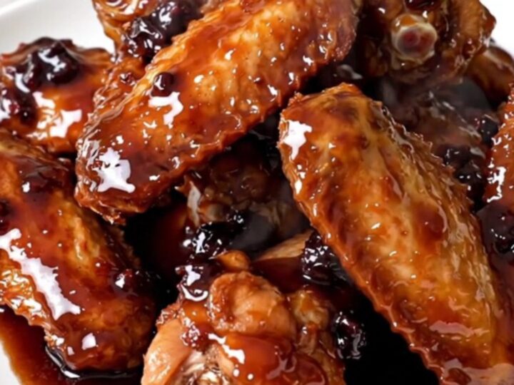 A top-down view of chicken wings cooking in a black slow cooker with blackberry sauce and jalapenos.