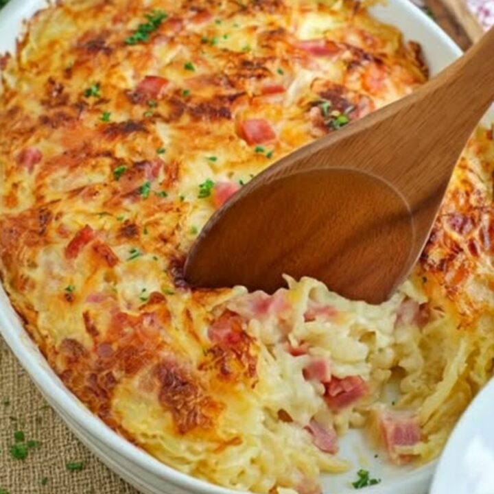 A warm oval white baking dish filled with cheesy ham and hash brown casserole, showing a golden-brown melted crust