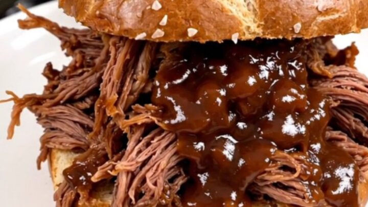A close-up of tender, glossy shredded beef piled high on a soft pretzel roll, steaming and ready to eat.