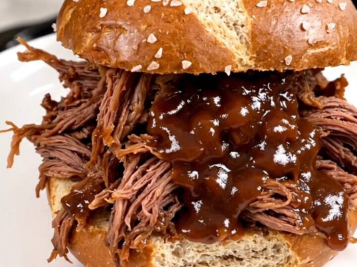 A close-up of tender, glossy shredded beef piled high on a soft pretzel roll, steaming and ready to eat.