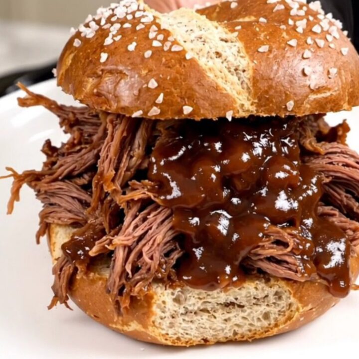 A close-up of tender, glossy shredded beef piled high on a soft pretzel roll, steaming and ready to eat.