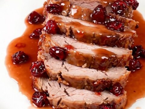 A beautifully sliced cranberry glazed pork loin served on a clean white plate with a glossy, deep red sauce.