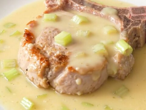 A close-up of a tender bone-in pork chop topped with a creamy, rich celery sauce on a white plate.