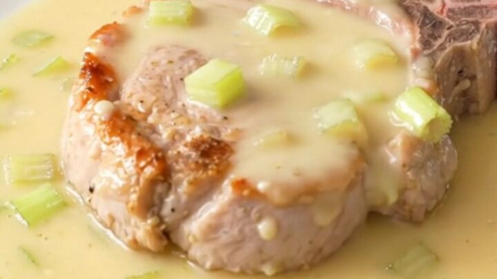 A close-up of a tender bone-in pork chop topped with a creamy, rich celery sauce on a white plate.