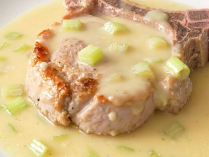 A close-up of a tender bone-in pork chop topped with a creamy, rich celery sauce on a white plate.