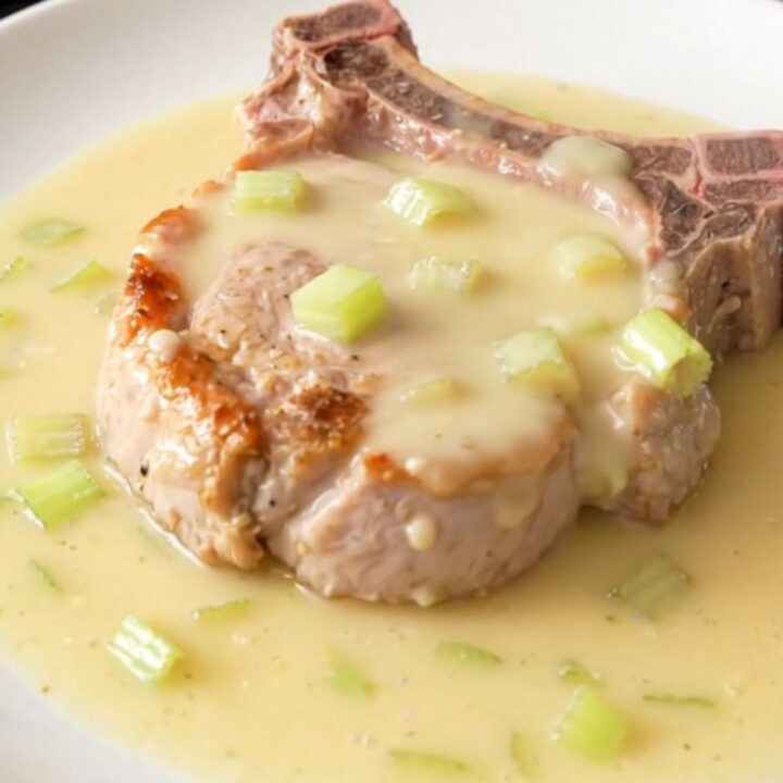 A close-up of a tender bone-in pork chop topped with a creamy, rich celery sauce on a white plate.