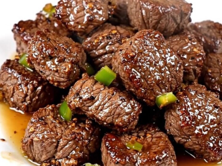 A close-up of glossy, slow-cooked Cajun steak bites with green bell peppers on a white plate.