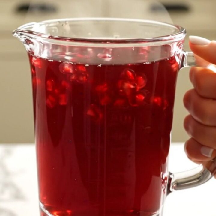 A warm glass mug filled with deep-red spiced pomegranate apple cider, garnished with a cinnamon stick.