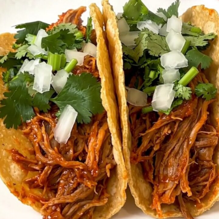 A plate of shredded pork birria tacos served on a white plate with fresh garnishes.