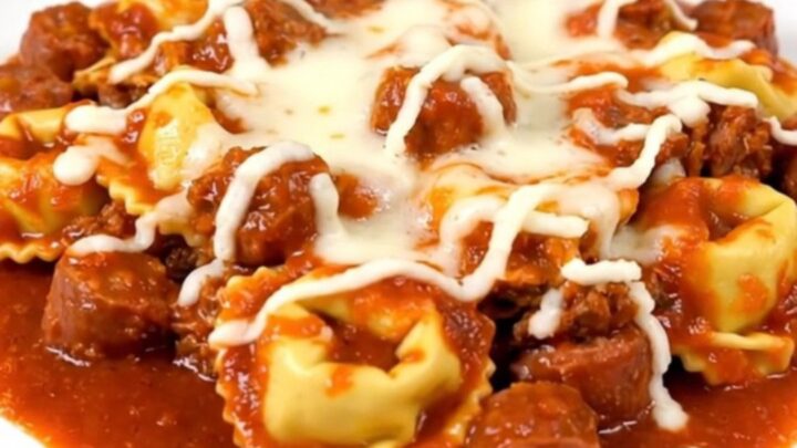 A top-down view of a slow cooker filled with browned Italian sausage, cheese tortellini, and rich tomato sauce.