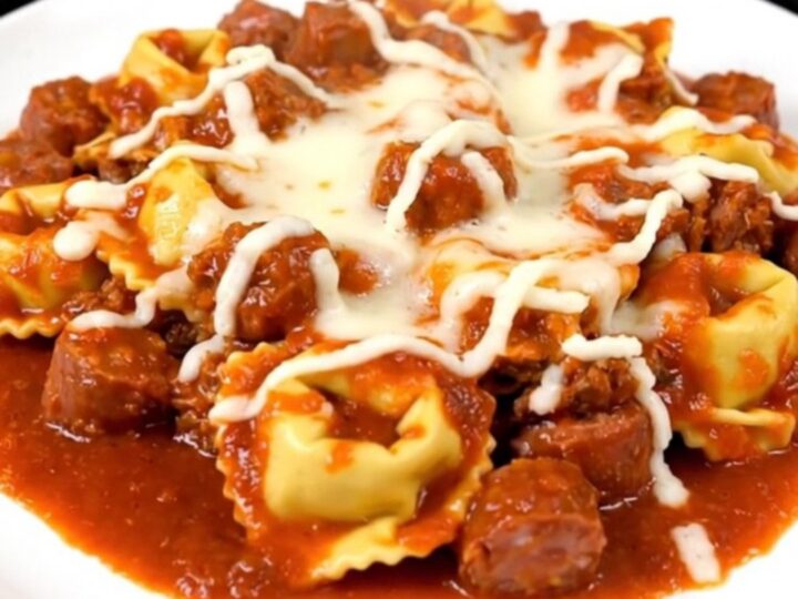 A top-down view of a slow cooker filled with browned Italian sausage, cheese tortellini, and rich tomato sauce.